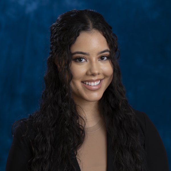 Portrait of a smiling woman with long dark wavy hair wearing a black blazer against a blue backdrop Portrait of a smiling woman with long dark wavy hair, wearing a black blazer against a blue backdrop.